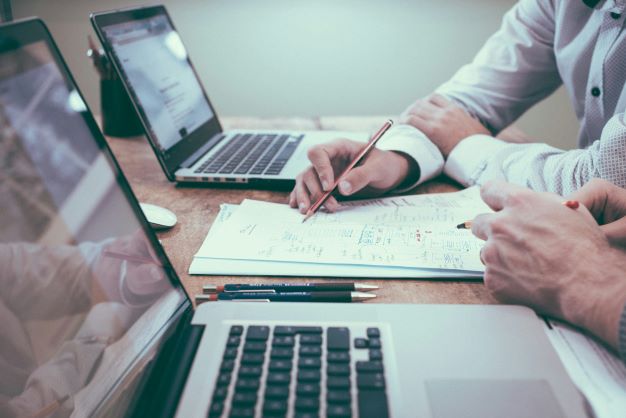 Two people working at a table with two laptops and writing on a paper document. Photo by Scott Graham on Unsplash.