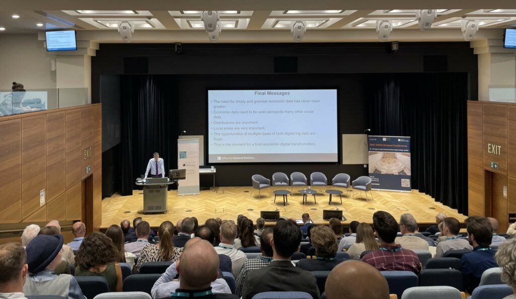 Conference room showing Sir Robert presenting in front of a projector screen to a room of people seated.