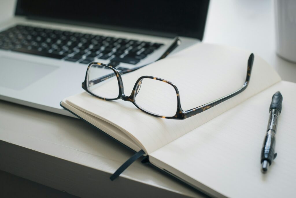 Glasses resting on a notebook next to a pen and open laptop. Photo by Trent Erwin on Unsplash