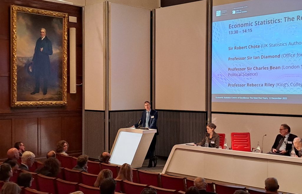 Sir Robert Chote speaking at the ESCoE conference. The audience is in front of him and behind him is a projection screen and other panel members.